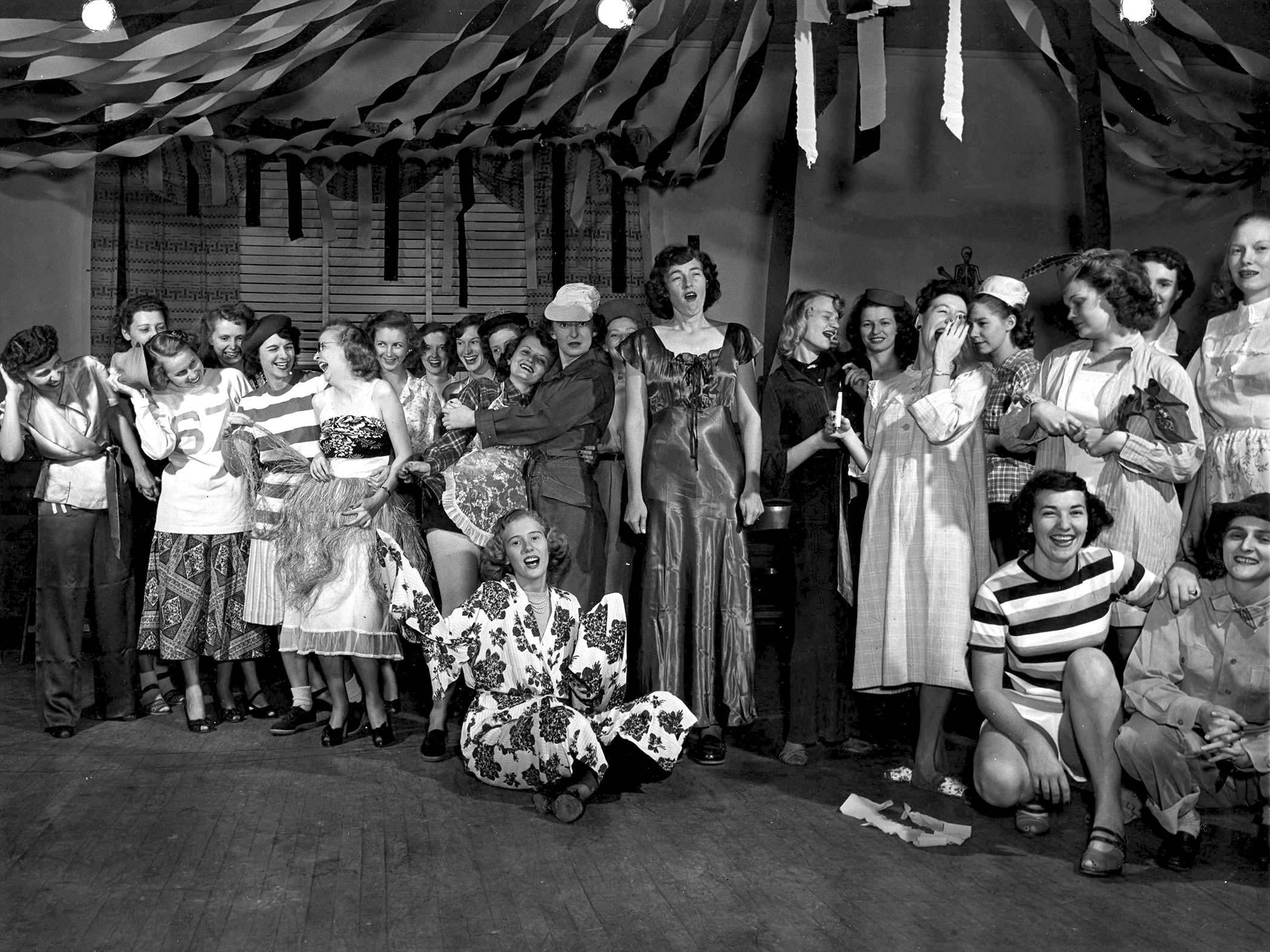 A large group of women wear costumes, perhaps for a skit or a party. Streamers hang from the ceiling. Circa 1947.