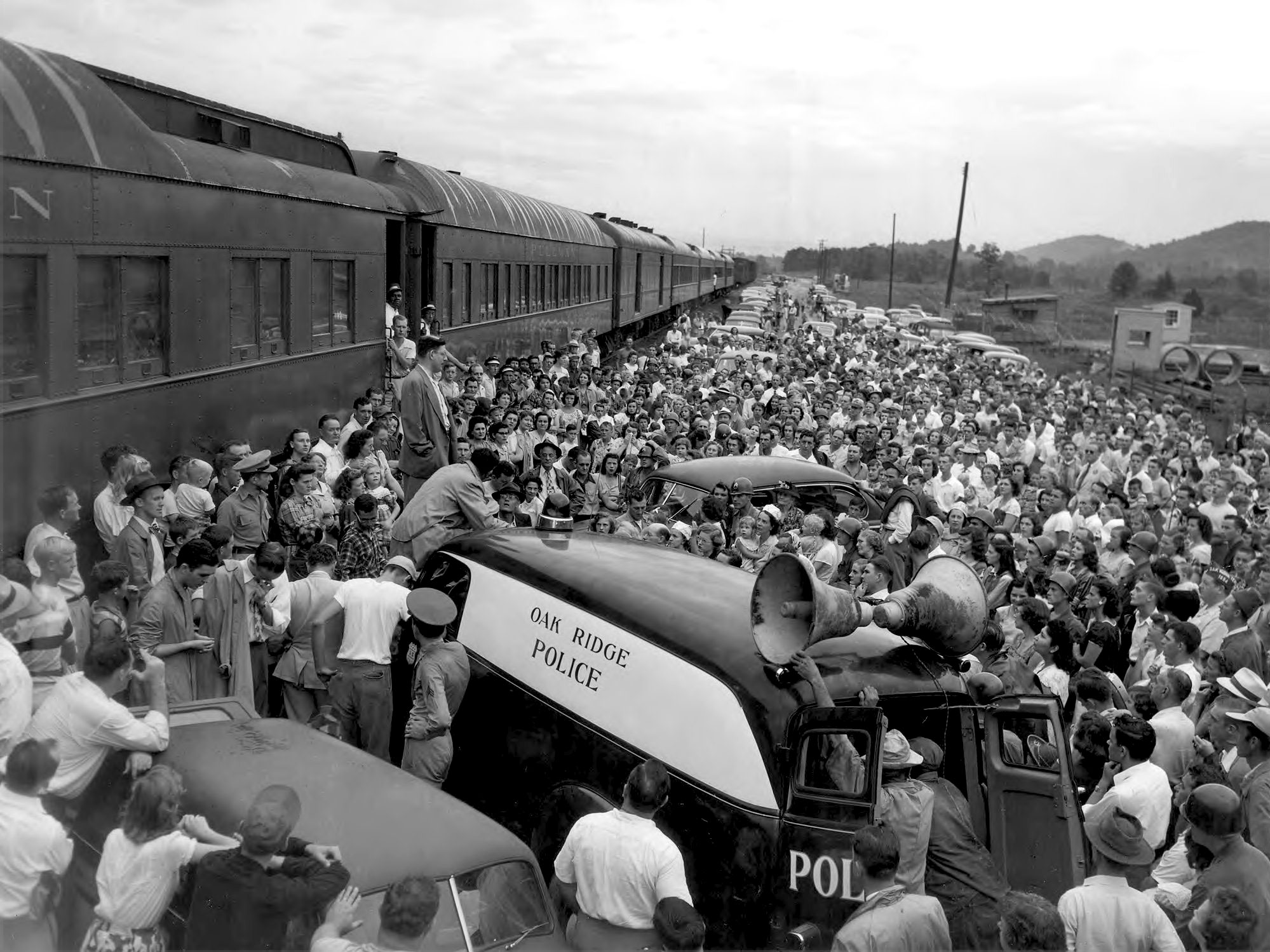 A wounded soldier speaks to a large crowd in front of a train. An automobile with a loudspeaker mounted on top is nearby and is labeled "Oak Ridge Police. Circa 1944.