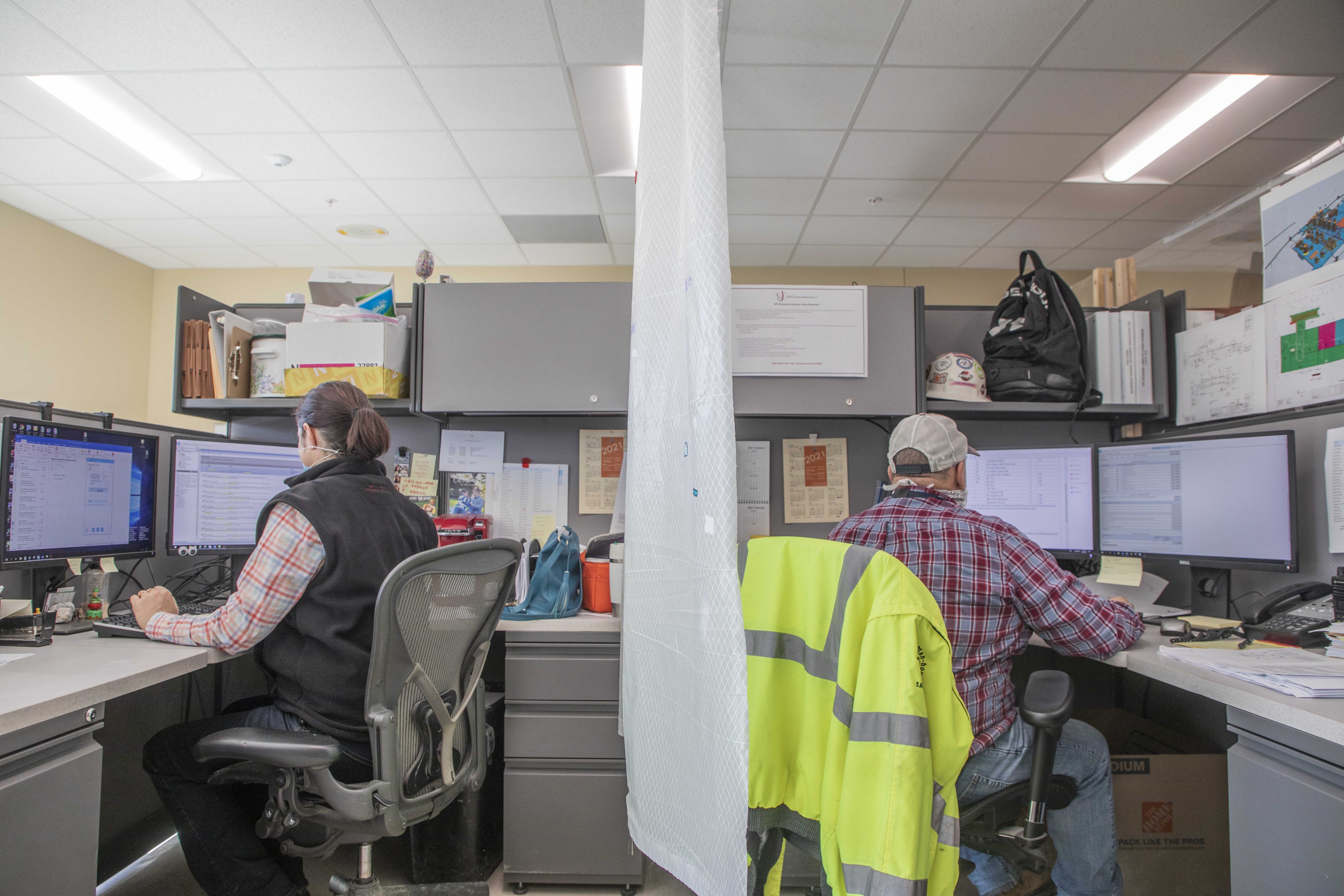 Two mission essential UPF employees work back to back with a COVID-19 safety partition between to maximize social distancing.