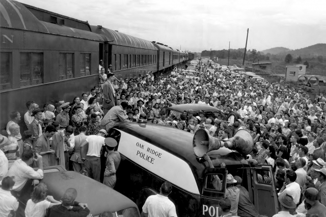 A wounded soldier speaks to a large crowd in front of a train. An automobile with a loudspeaker mounted on top is nearby and is labeled "Oak Ridge Police. Circa 1944.
