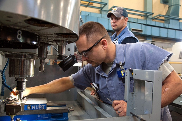 Apprentice machinist John Bryant, foreground, uses dial calipers to measure a feature of a part while his twin brother Jeff Bryant, also an apprentice machinist, looks on. 